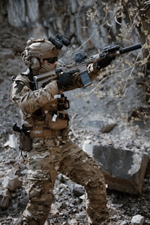A person wearing full tactical military gear is holding a rifle equipped with a scope and suppressor. The individual is dressed in camouflage clothing and appears to be standing in a rocky, outdoor setting with a focused stance.