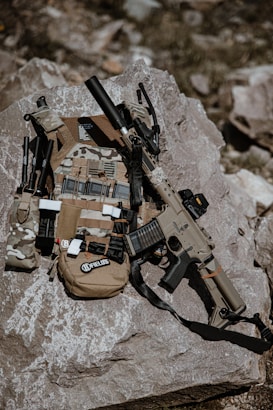 Tactical gear including a rifle and a camouflage vest placed on a large rock. The rifle is equipped with accessories like a scope and a suppressor. Various attachments and pouches are visible on the vest.