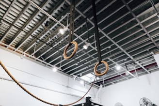 A pair of wooden gymnastics rings suspended from the ceiling by black straps in a gym setting. The ceiling is exposed, showing metal beams and industrial lighting fixtures.