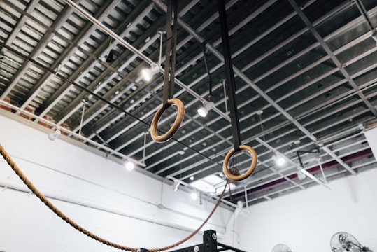 A pair of wooden gymnastics rings suspended from the ceiling by black straps in a gym setting. The ceiling is exposed, showing metal beams and industrial lighting fixtures.