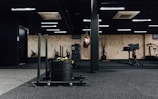 A gym interior featuring various exercise equipment including a sled with weights, a punching bag, and other fitness machines. The space has black flooring with a mix of wooden and black walls, ceiling lights, and an air conditioning unit visible.