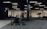 A gym interior featuring various exercise equipment including a sled with weights, a punching bag, and other fitness machines. The space has black flooring with a mix of wooden and black walls, ceiling lights, and an air conditioning unit visible.