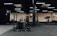 A gym interior featuring various exercise equipment including a sled with weights, a punching bag, and other fitness machines. The space has black flooring with a mix of wooden and black walls, ceiling lights, and an air conditioning unit visible.