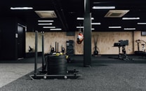 A gym interior featuring various exercise equipment including a sled with weights, a punching bag, and other fitness machines. The space has black flooring with a mix of wooden and black walls, ceiling lights, and an air conditioning unit visible.