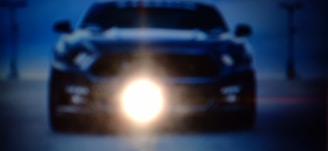 A blurred and somewhat dark image depicting the front of a car with headlights shining prominently in the center. The surrounding environment appears to be outdoor, possibly with an urban backdrop. The lighting creates a dramatic effect.