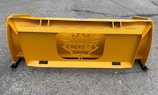 A large, industrial yellow snowplow blade with a central cutout featuring a maple leaf design and the letters 'CM'. It is placed on a gray, rough asphalt surface with faint markings.