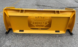 A large, industrial yellow snowplow blade with a central cutout featuring a maple leaf design and the letters 'CM'. It is placed on a gray, rough asphalt surface with faint markings.