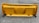 A large, industrial yellow snowplow blade with a central cutout featuring a maple leaf design and the letters 'CM'. It is placed on a gray, rough asphalt surface with faint markings.