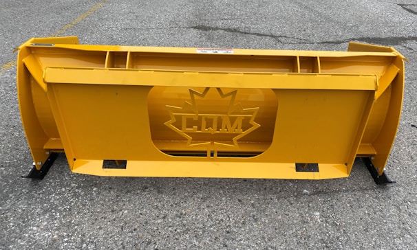 A large, industrial yellow snowplow blade with a central cutout featuring a maple leaf design and the letters 'CM'. It is placed on a gray, rough asphalt surface with faint markings.