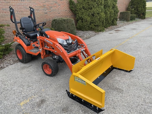 An orange tractor with a yellow attachment is parked on a paved surface near a brick wall and some greenery. The tractor appears compact and suitable for landscaping or construction work.