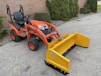 An orange tractor with a yellow attachment is parked on a paved surface near a brick wall and some greenery. The tractor appears compact and suitable for landscaping or construction work.