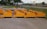 Several bright yellow industrial attachments are arranged in a row on a paved surface. They are positioned near a trailer and a chain-link fence, with a road and grassy area visible in the background under a cloudy sky.
