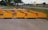 Several bright yellow industrial attachments are arranged in a row on a paved surface. They are positioned near a trailer and a chain-link fence, with a road and grassy area visible in the background under a cloudy sky.