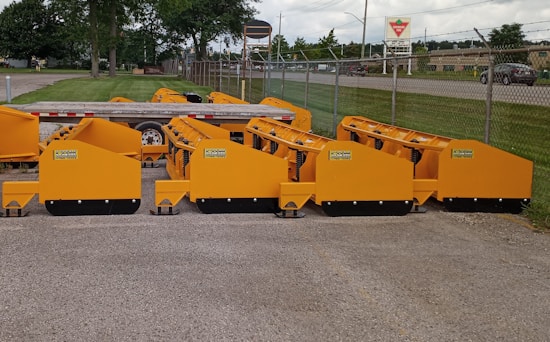 Several bright yellow industrial attachments are arranged in a row on a paved surface. They are positioned near a trailer and a chain-link fence, with a road and grassy area visible in the background under a cloudy sky.
