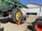 A large agricultural machine, likely a piece of farm equipment, is being operated in an outdoor setting. A green tractor with sizable yellow wheels is attached to a mechanical arm. The machinery is positioned on a gravel surface near a large white building with a garage door.