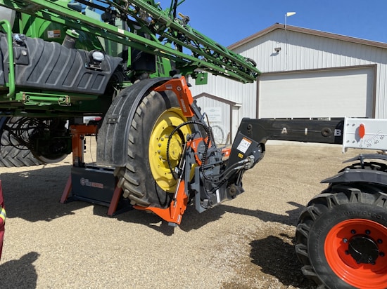 A large agricultural machine, likely a piece of farm equipment, is being operated in an outdoor setting. A green tractor with sizable yellow wheels is attached to a mechanical arm. The machinery is positioned on a gravel surface near a large white building with a garage door.