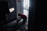A person kneels on the floor in a dimly lit hallway near a door, working with wires or equipment. The hallway has a patterned carpet, and there are scattered tools and materials. Light enters from a window in the background, partially illuminating the scene.
