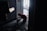 A person kneels on the floor in a dimly lit hallway near a door, working with wires or equipment. The hallway has a patterned carpet, and there are scattered tools and materials. Light enters from a window in the background, partially illuminating the scene.