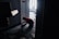 A person kneels on the floor in a dimly lit hallway near a door, working with wires or equipment. The hallway has a patterned carpet, and there are scattered tools and materials. Light enters from a window in the background, partially illuminating the scene.