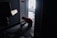 A person kneels on the floor in a dimly lit hallway near a door, working with wires or equipment. The hallway has a patterned carpet, and there are scattered tools and materials. Light enters from a window in the background, partially illuminating the scene.