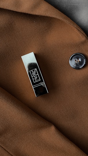 A metallic accessory with a sleek, reflective surface is clipped onto a brown fabric, likely part of a coat. There is also a black button with a glossy finish visible on the fabric.