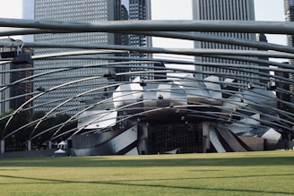 A large, futuristic architectural structure with curved metal elements dominates the scene, surrounded by skyscrapers. The design appears intricate and modern, with a spacious green lawn in the foreground. The structure is part of an outdoor concert venue with overhead lighting and support beams.