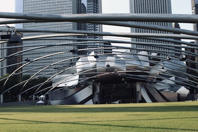A large, futuristic architectural structure with curved metal elements dominates the scene, surrounded by skyscrapers. The design appears intricate and modern, with a spacious green lawn in the foreground. The structure is part of an outdoor concert venue with overhead lighting and support beams.