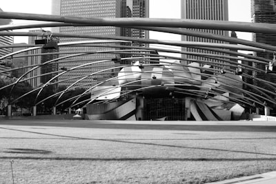 An architectural structure with a modern design featuring sweeping curves and metallic surfaces is positioned against a backdrop of tall, rectangular city buildings. A network of metal beams arches overhead, extending from one side of the image to the other. The foreground is a broad grassy area, casting subtle shadows due to the structures above.