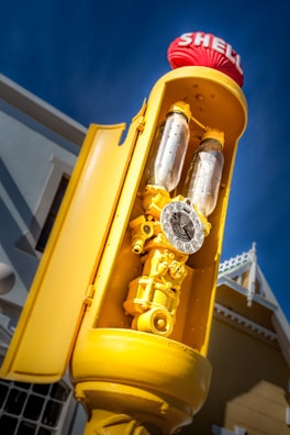 An old-fashioned gas pump painted in bright yellow with a red Shell logo on top. The pump contains two glass cylinders and a vintage-style dial with numbers. The background includes a clear blue sky and part of a building with a yellow facade and white trim.