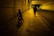 A person and a child are moving through a dimly lit urban tunnel. The child is riding a small bicycle, while the adult walks beside them. The tunnel is lined with tiles and features overhead lighting casting a warm yellow glow. There is a road beside the walkway with a parked car visible in the distance.