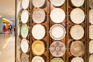 A decorative display of various plates featuring intricate designs and textures, mounted on a wall against a background with soft lighting. The plates appear to be made of materials like glass and porcelain with a range of patterns, including floral motifs and geometric shapes. The setting suggests an upscale retail or gallery environment.