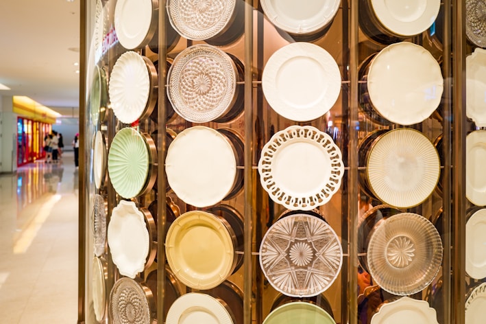 A decorative display of various plates featuring intricate designs and textures, mounted on a wall against a background with soft lighting. The plates appear to be made of materials like glass and porcelain with a range of patterns, including floral motifs and geometric shapes. The setting suggests an upscale retail or gallery environment.