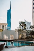 A pool area surrounded by a concrete deck and metal railings, with lush plants and palm trees in the background. In the distance, modern high-rise buildings including a distinctive blue skyscraper with a pointed top are visible against a clear sky.