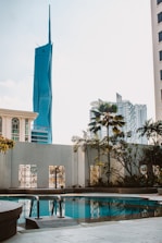 A pool area surrounded by a concrete deck and metal railings, with lush plants and palm trees in the background. In the distance, modern high-rise buildings including a distinctive blue skyscraper with a pointed top are visible against a clear sky.