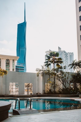 A pool area surrounded by a concrete deck and metal railings, with lush plants and palm trees in the background. In the distance, modern high-rise buildings including a distinctive blue skyscraper with a pointed top are visible against a clear sky.