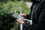 A person wearing a dark jacket is holding a drone controller with a smartphone attached. The background includes greenery and a blurred road, suggesting an outdoor setting.
