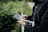 A person wearing a dark jacket is holding a drone controller with a smartphone attached. The background includes greenery and a blurred road, suggesting an outdoor setting.