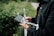 A person wearing a dark jacket is holding a drone controller with a smartphone attached. The background includes greenery and a blurred road, suggesting an outdoor setting.