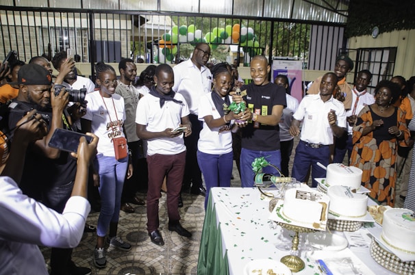 A group of people gathered around a table with multiple white cakes decorated with green ribbons and text. The scene includes photographers capturing the event, and some people are dressed in casual attire, while others wear uniforms. Balloons in green, orange, and white colors are seen in the background, indicating a festive occasion.