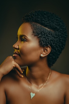 A profile view of a woman with short curly hair, wearing pearl earrings and a heart-shaped necklace. She rests her chin on her hand and gazes thoughtfully into the distance. The lighting casts a warm glow on her face, highlighting her features.