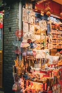 A vibrant market stall displays an array of traditional Asian items, including decorative fans, umbrellas, plates, and small toys. The stall is brightly lit and colorful, with intricate patterns and designs on many of the products.