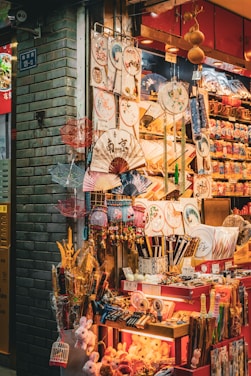 A vibrant market stall displays an array of traditional Asian items, including decorative fans, umbrellas, plates, and small toys. The stall is brightly lit and colorful, with intricate patterns and designs on many of the products.