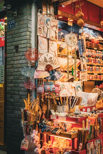 A vibrant market stall displays an array of traditional Asian items, including decorative fans, umbrellas, plates, and small toys. The stall is brightly lit and colorful, with intricate patterns and designs on many of the products.