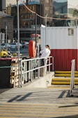 A person stands on a platform next to a large red and white container. They are leaning against a railing, and the area is surrounded by various city elements, including parked cars, a tall building, and a yellow stairway. A lifebuoy is attached to a pole near the person.