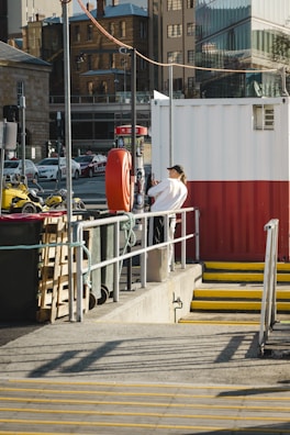 A person stands on a platform next to a large red and white container. They are leaning against a railing, and the area is surrounded by various city elements, including parked cars, a tall building, and a yellow stairway. A lifebuoy is attached to a pole near the person.