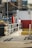 A person stands on a platform next to a large red and white container. They are leaning against a railing, and the area is surrounded by various city elements, including parked cars, a tall building, and a yellow stairway. A lifebuoy is attached to a pole near the person.