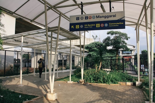 A covered walkway with metallic pillars and a roof structure. A sign overhead indicates directions to KA Bandara Airport Train and Commuterline. There is a person walking under the covered area, and greenery including trees and plants on the side. Buildings and a fence are visible in the background.