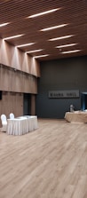 A spacious, modern hall with wooden flooring and walls featuring vertical wooden slats, illuminated by recessed lighting on the ceiling. A table draped with a white cloth has two chairs next to it, and there is a covered table with some catering equipment in the background.