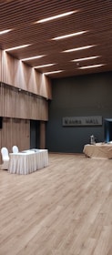 A spacious, modern hall with wooden flooring and walls featuring vertical wooden slats, illuminated by recessed lighting on the ceiling. A table draped with a white cloth has two chairs next to it, and there is a covered table with some catering equipment in the background.