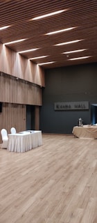 A spacious, modern hall with wooden flooring and walls featuring vertical wooden slats, illuminated by recessed lighting on the ceiling. A table draped with a white cloth has two chairs next to it, and there is a covered table with some catering equipment in the background.